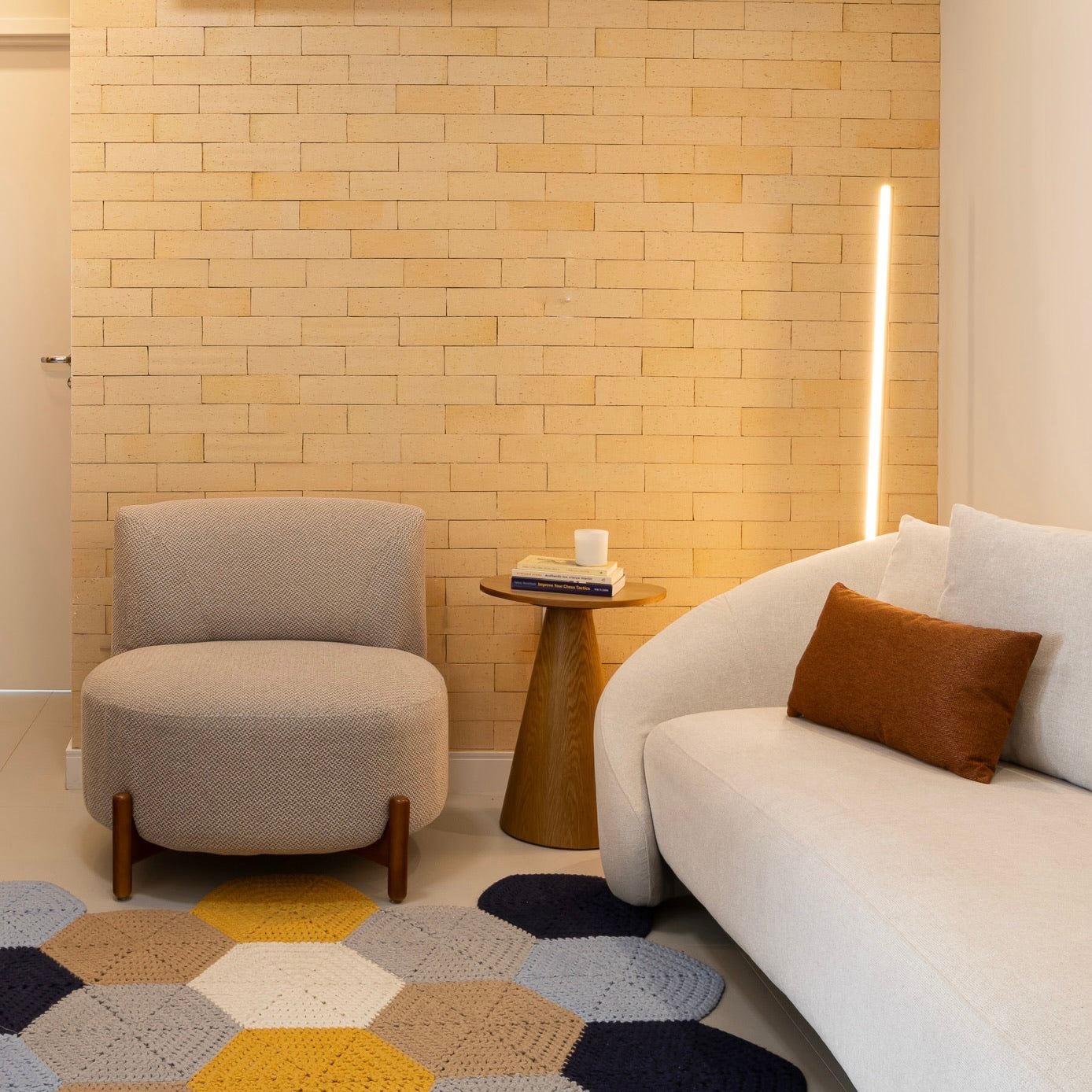 Sala de estar com revestimento de tijolinho decorativo Quarel Rustic Sand na parede, sofá branco arredondado, poltrona bege, tapete de crochê hexagonal e luminária vertical LED